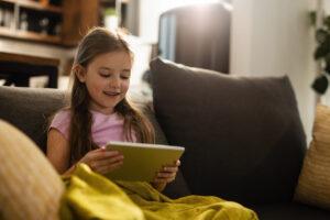 happy little girl watching cartoons on touchpad while sitting on the sofa scaled